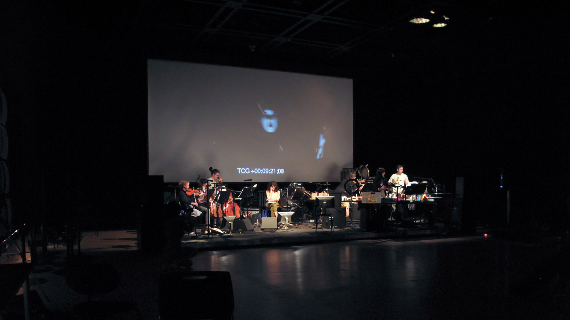 A small pit orchestra set up on a black stage un a large screen projecting "TCG +00:09:21;08" in white block letters.  