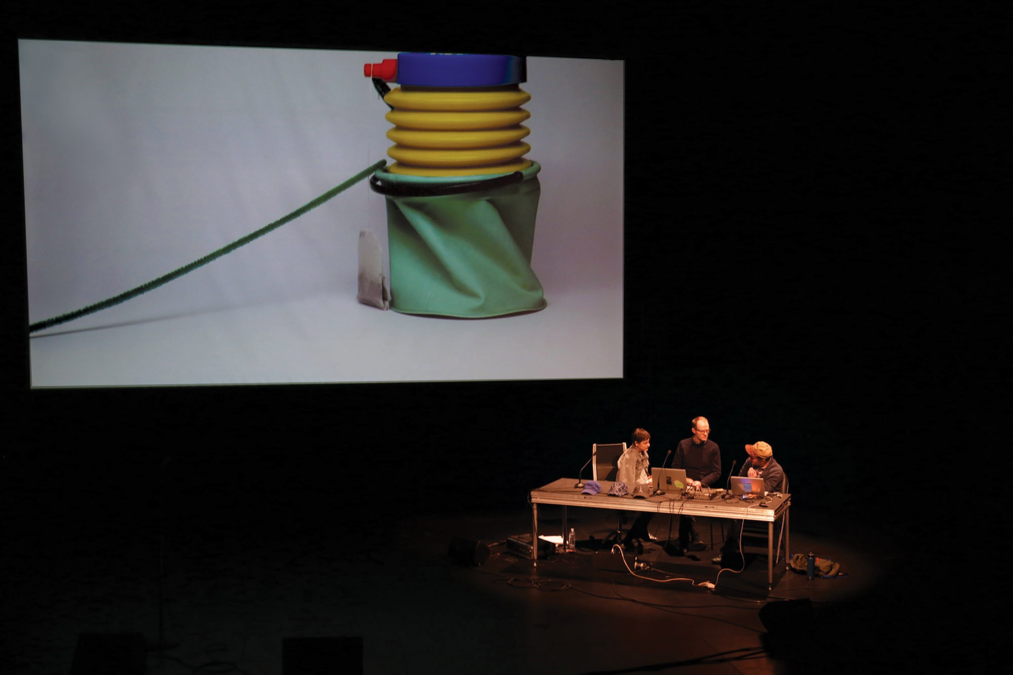 Three people sitting on stage behind a cluttered desk in front of a large screen projecting and image of a green yellow blue sculpture 