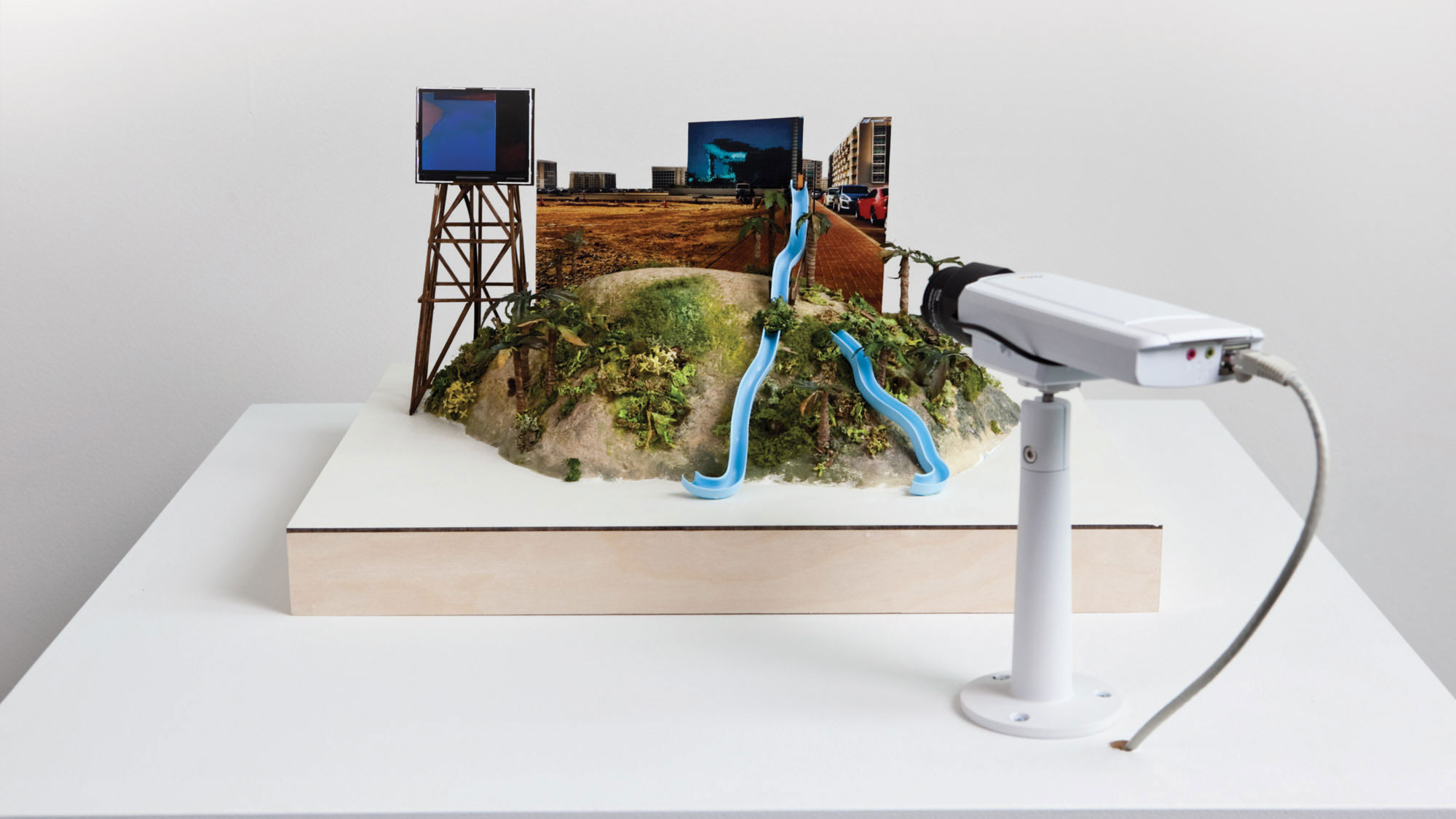 A miniature model of a waterslide on a hill on a white pedestal with a cct camera pointing at it