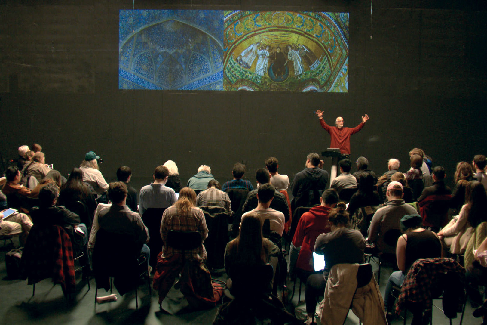 Johannes Goebel giving a talk to a small crowd in a black box studio. 