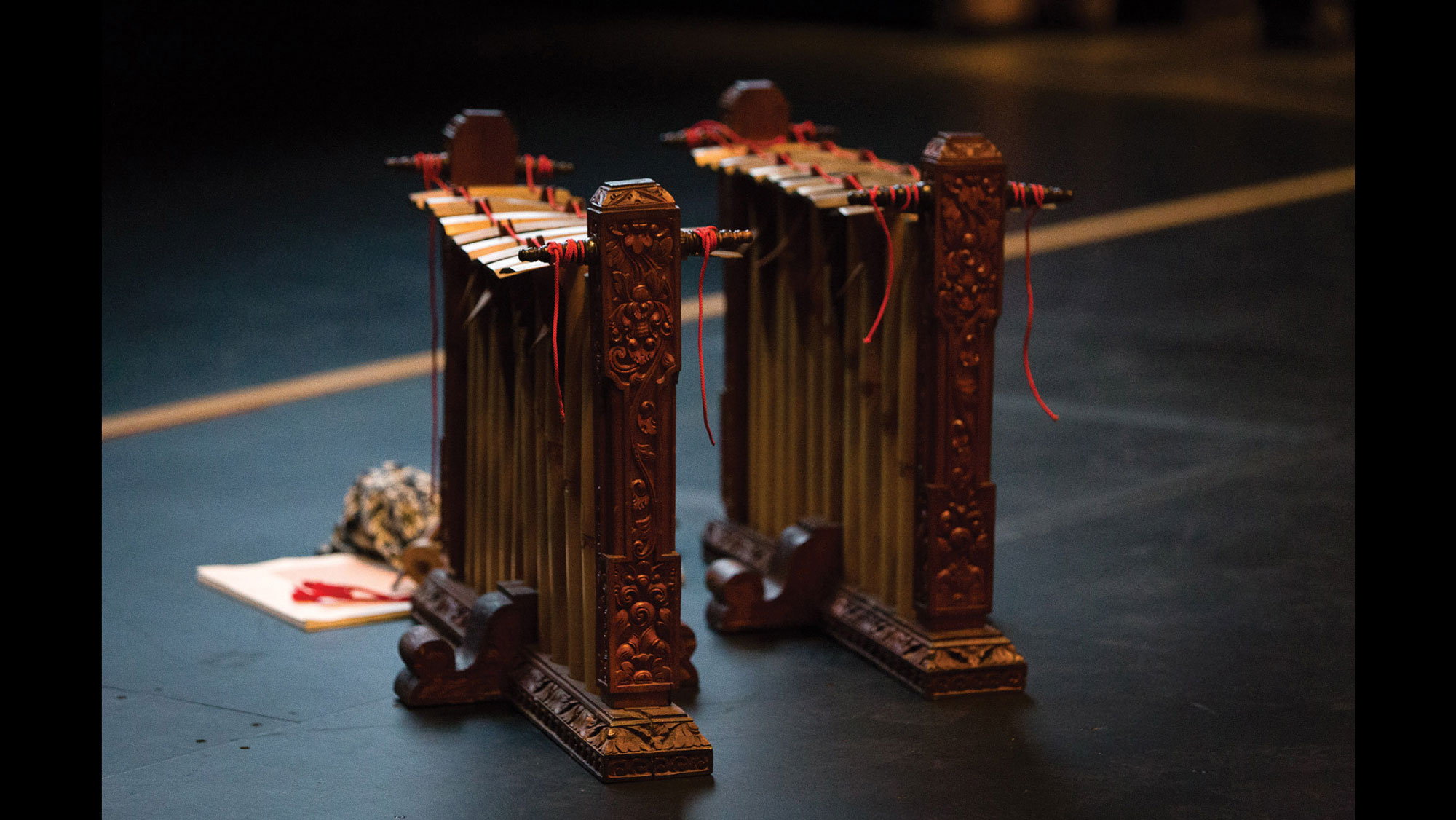 Two balinese instruments made of ornately carved wood held together by red string standing on a black floor. A small bouquet of flowers sits in front of them 