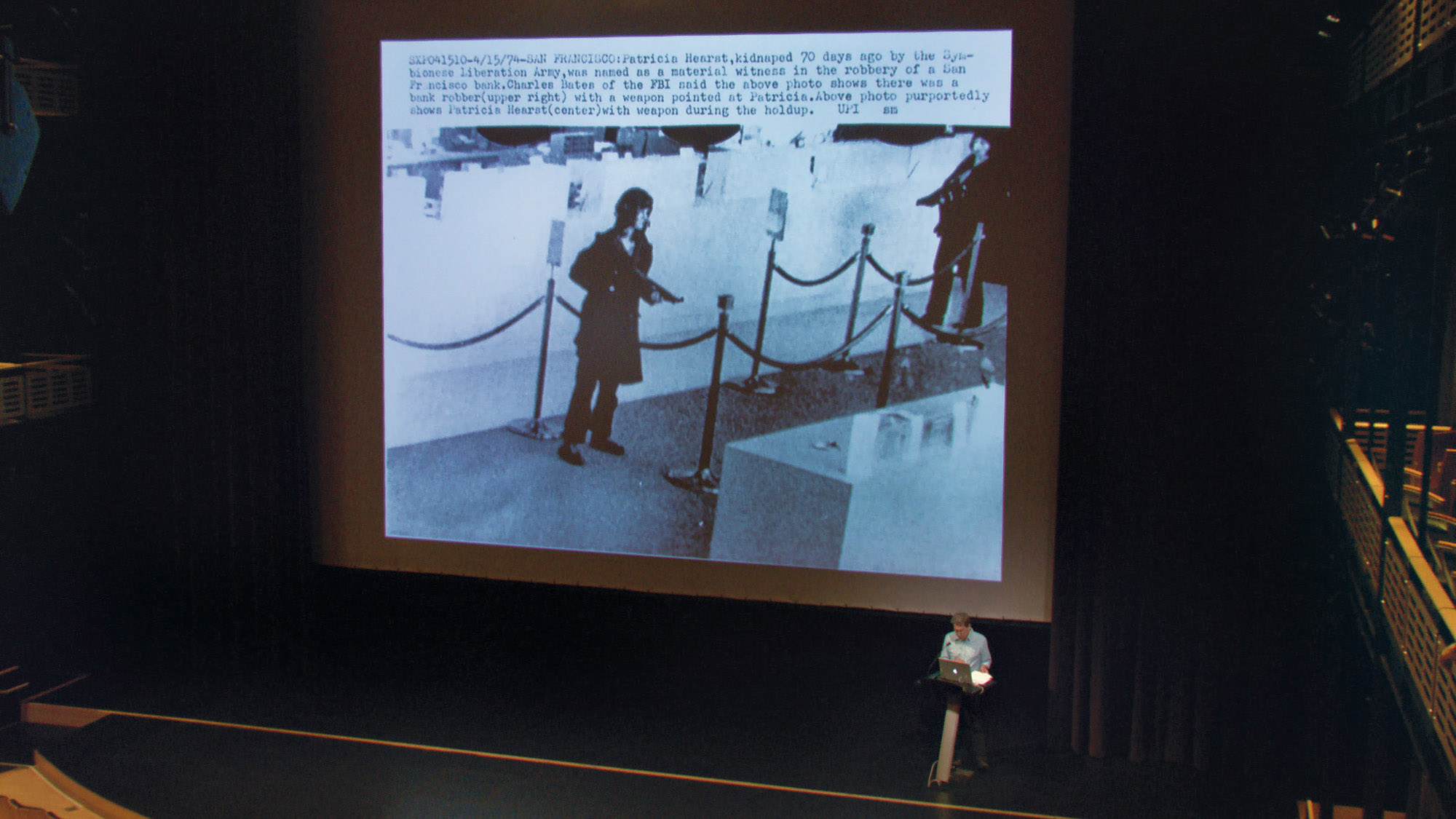 Thomas Keenan on stage in front of a large projection of an image of Patricia Hearst robbing a bank 