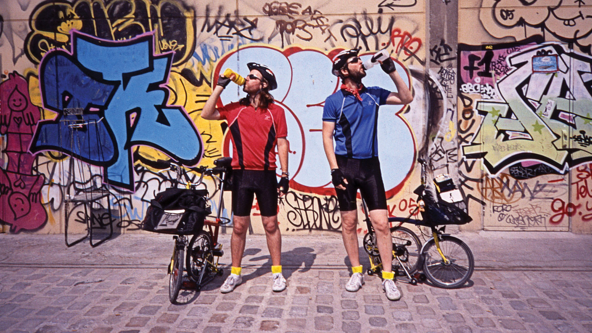 Two male bicyclists, one wearing red the other blue drinking from their water bottles with a wall of graffiti in the background. 