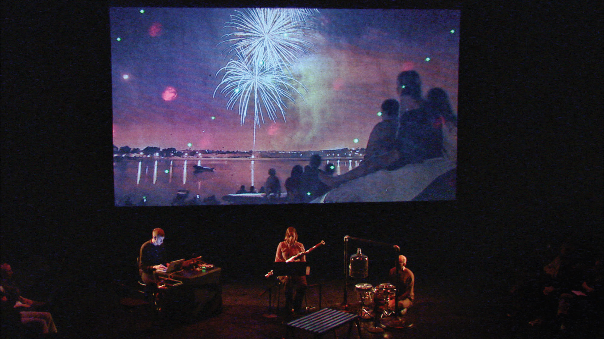 A group of three musicians playing on a dark stage lit by a projection of fireworks on a screen behind them. 