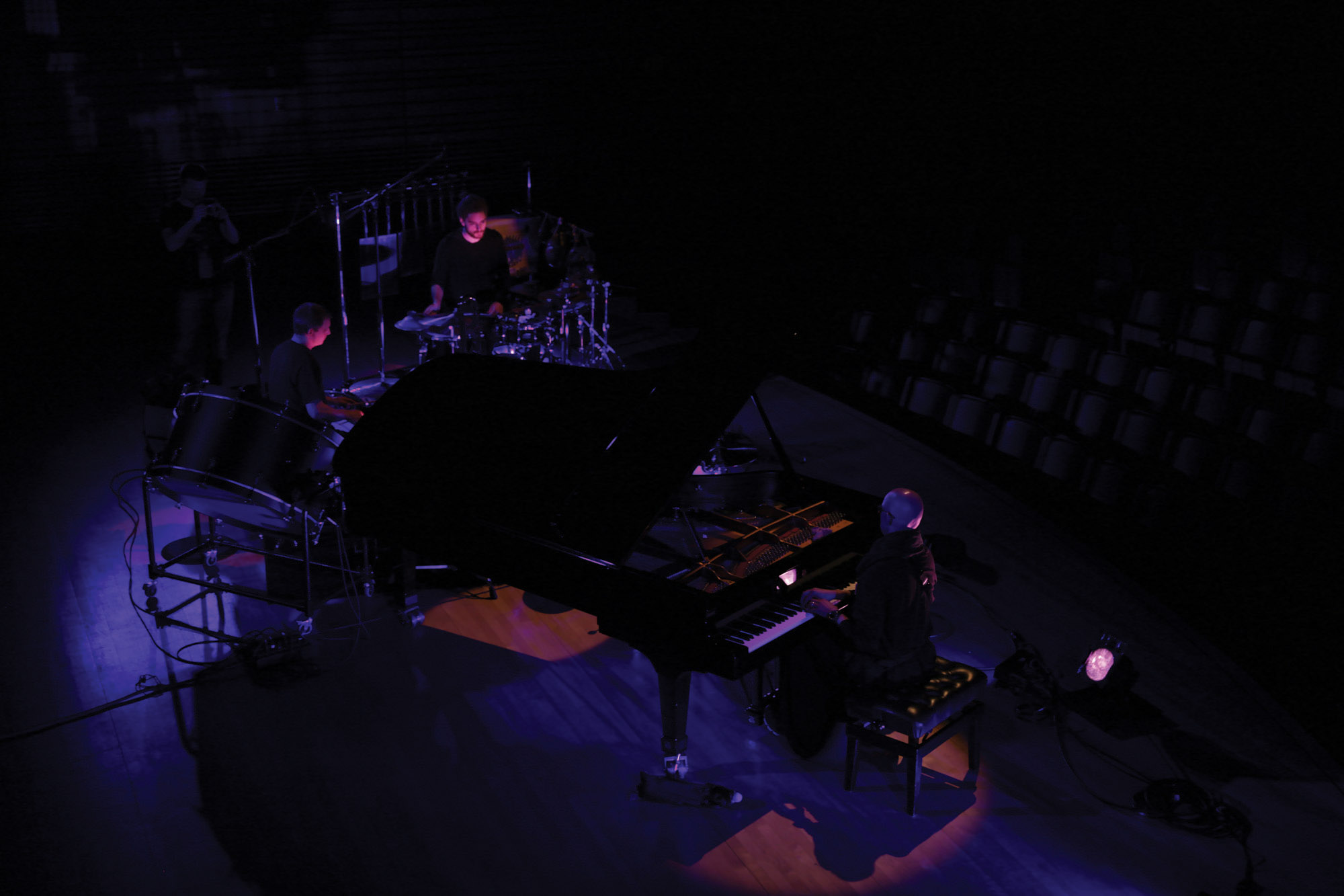 A small ensemble of three men, one playing piano and two on percussion, playing on the concert hall stage washed in blue/purple light. 