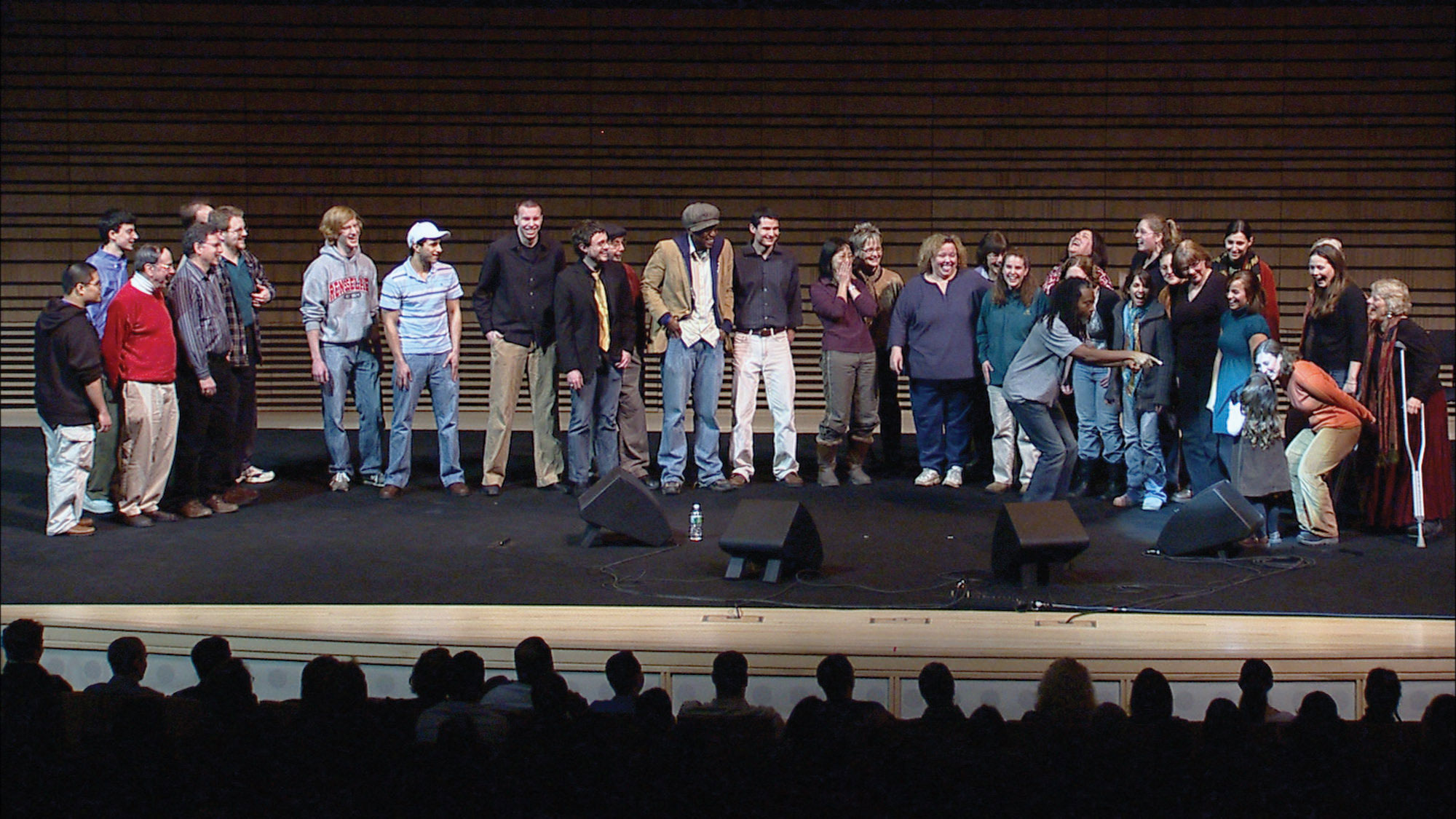 An assorted group of about 30 people standing on the concert hall stage in front of an audience. 