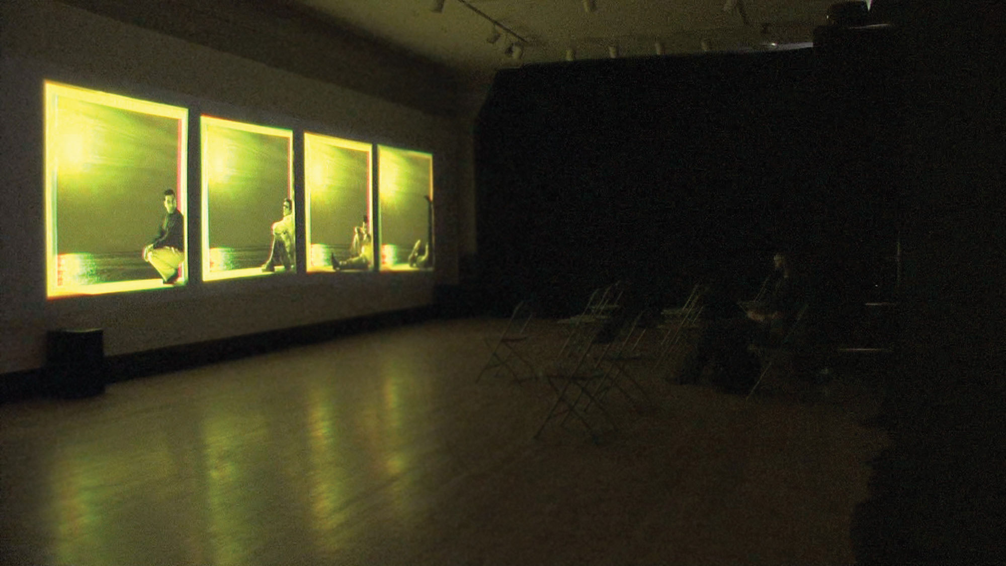 Four yellow projections of four people sitting against the wall within the box of each projection on a gallery wall. 