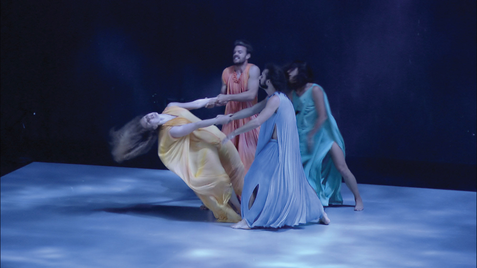 Four dancers wearing pastel colored flowing costumes standing in a circle. A woman in yellow throws herself back dramatically as the others hold her arms. 