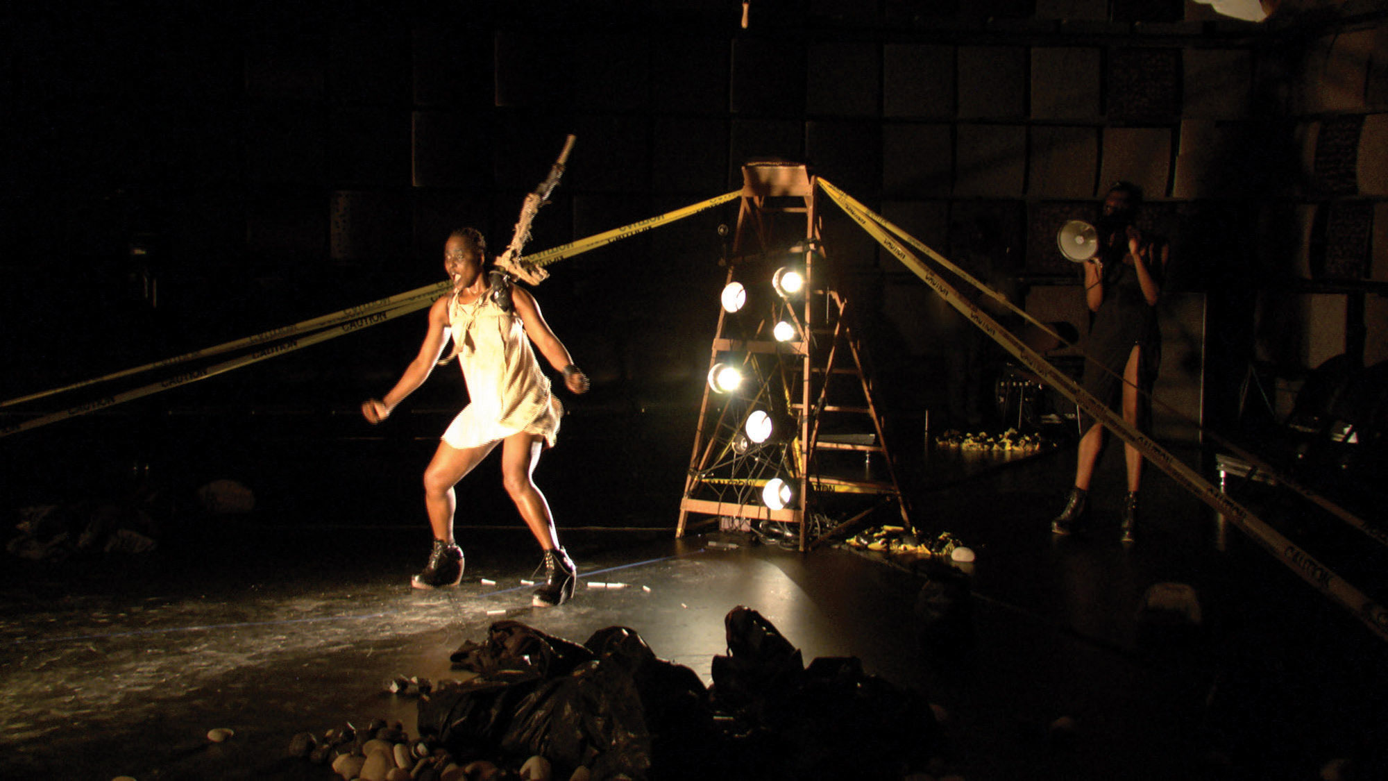 A Zimbabwean woman on a harness dancing in black box studio amongst piles of trash bags, caution tape, and a ladder with stage lights placed on the steps. 
