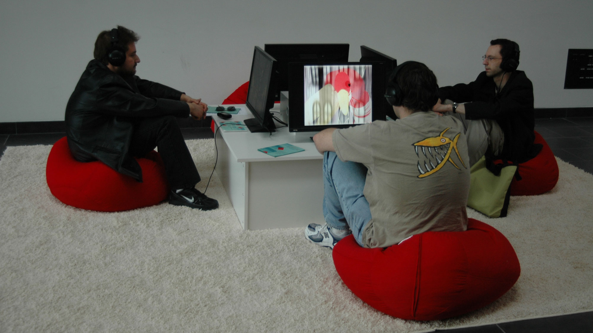 Three men seated on small red bean bag chairs on a white shag rug watching individual screens showing abstract images. 