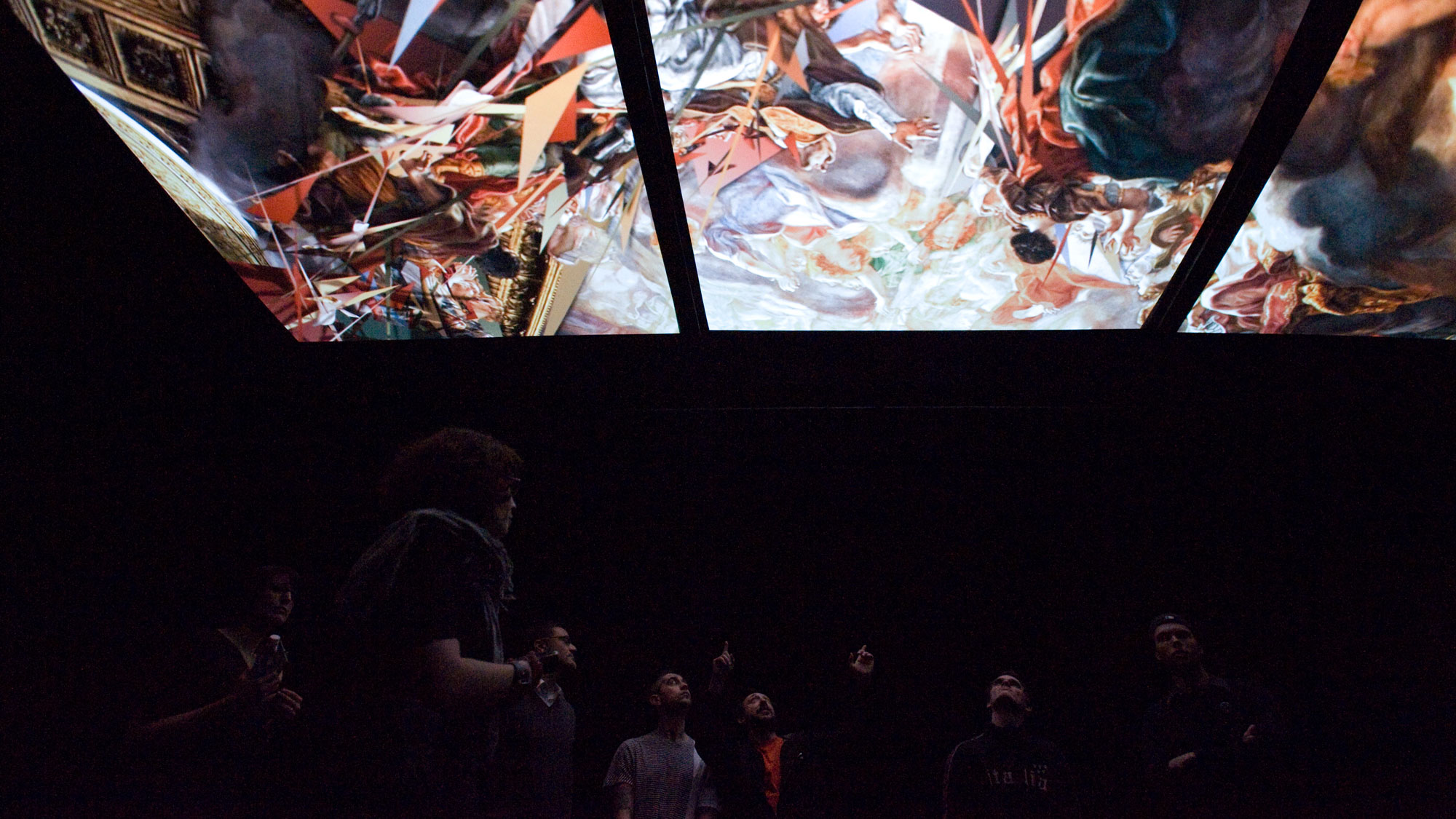 Three screens suspended from the ceiling of a dark blackbox  theater. A small crowd is gathered beneath them, looking up. 