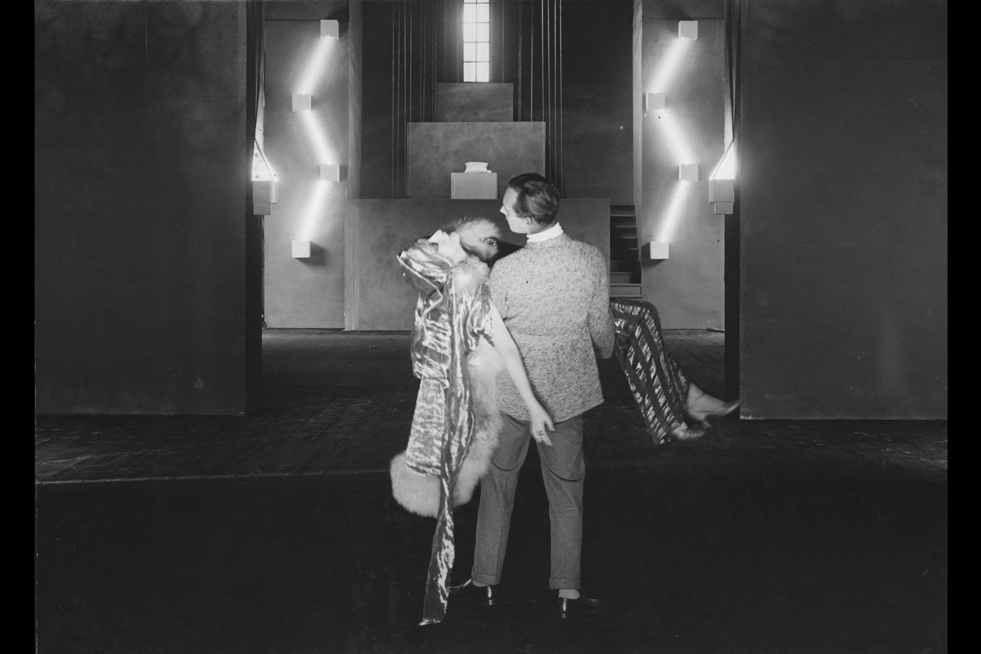 a man holding a woman in a dress in the 1920s in front of an art deco set. 