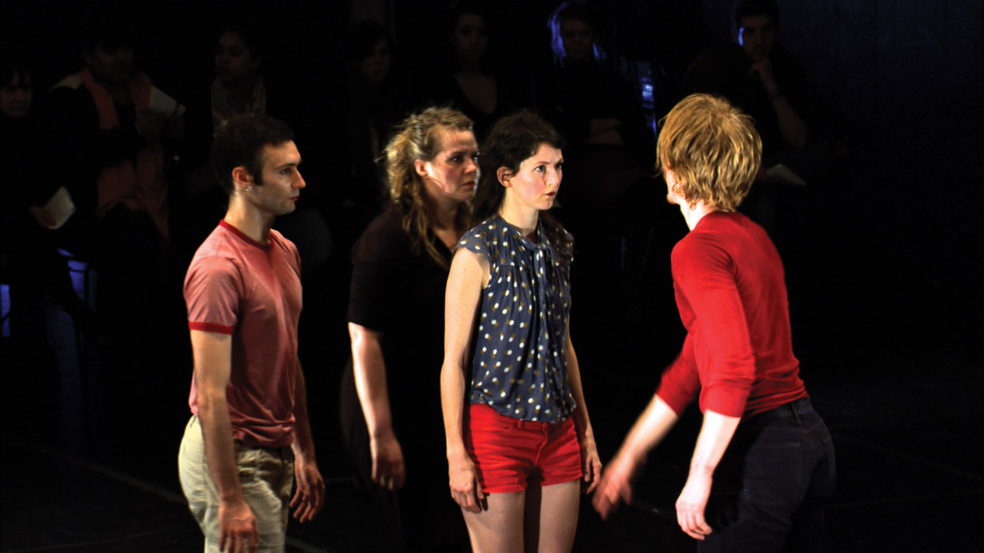 Four dancers dressed in street clothes with red accents on a dark stage. One person wearing a red long sleeved shirt with the back to the viewer, reaches towards another dancer wearing red shorts as the two others look on. 