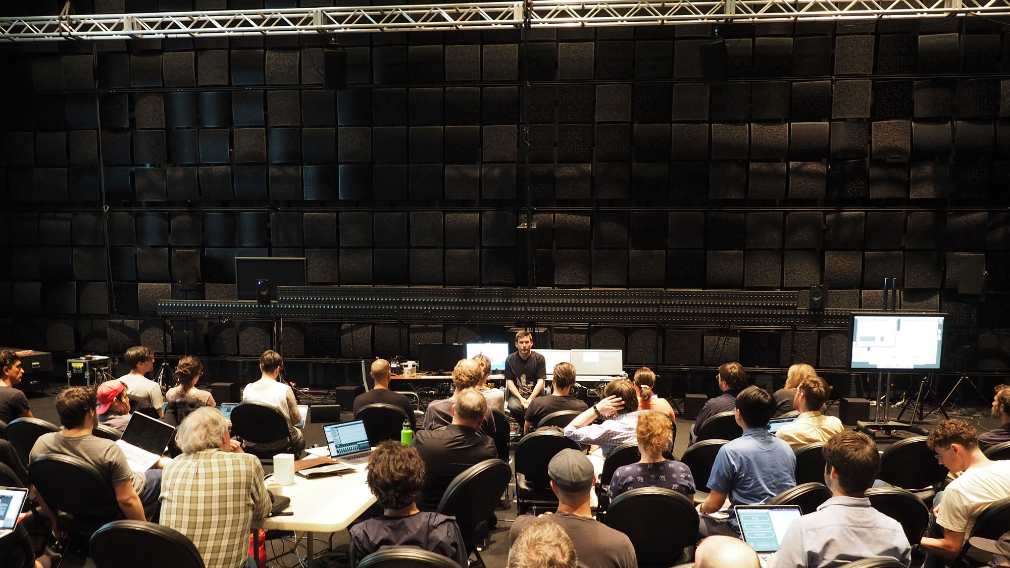 A white male wearing a black t-shirt and jeans sitting on a stool in a room with various computer set ups addressing a packed crowd, attentively looking on in a black box studio. 