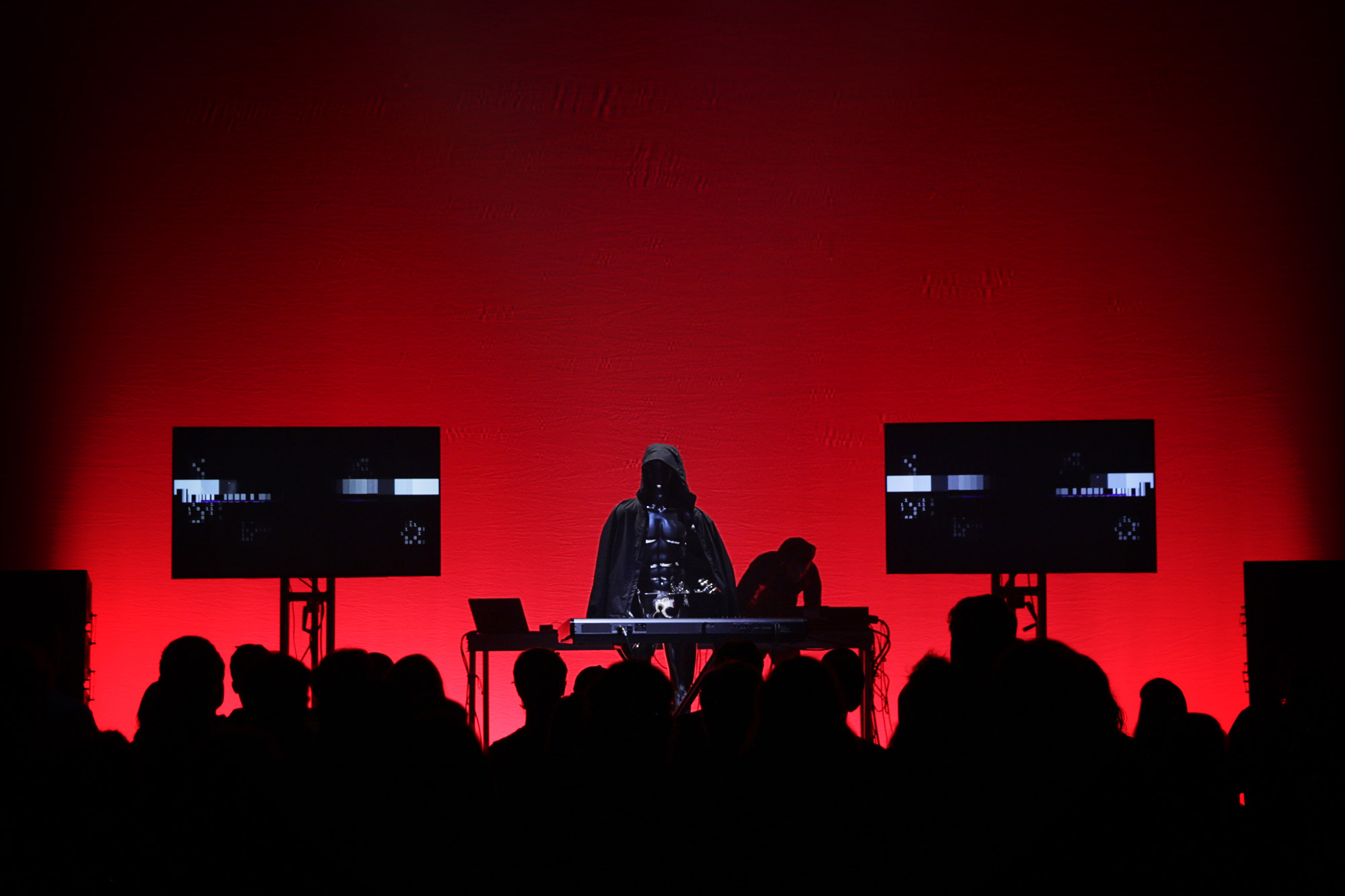 A figure in a black cloak on stage in front of a wall of red light, performing for a silhouetted crowd. 