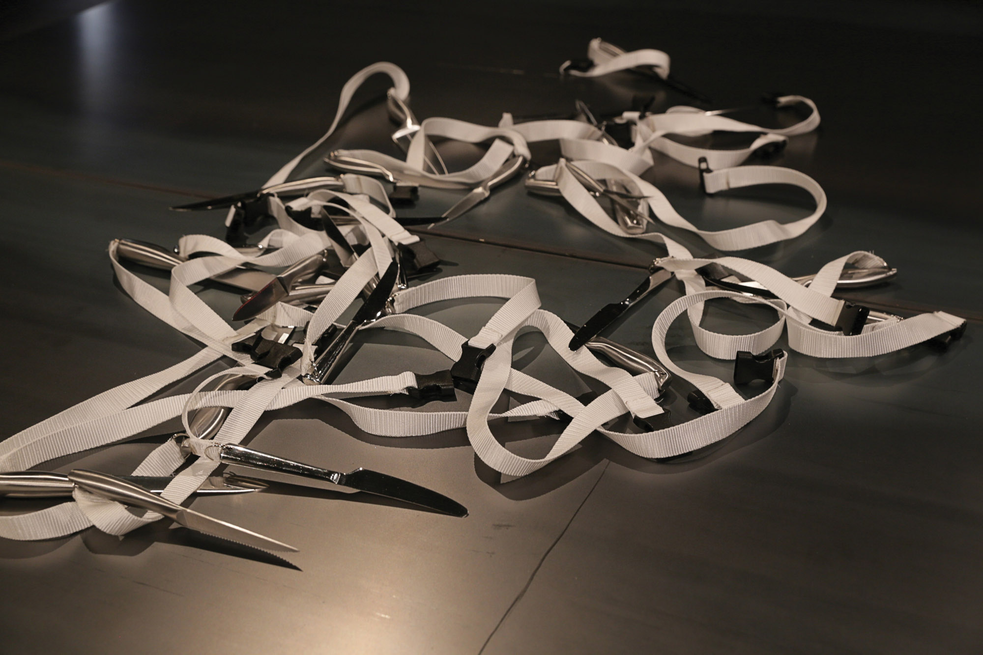 A white strap tangled with butter knives attached lying on a black floor.