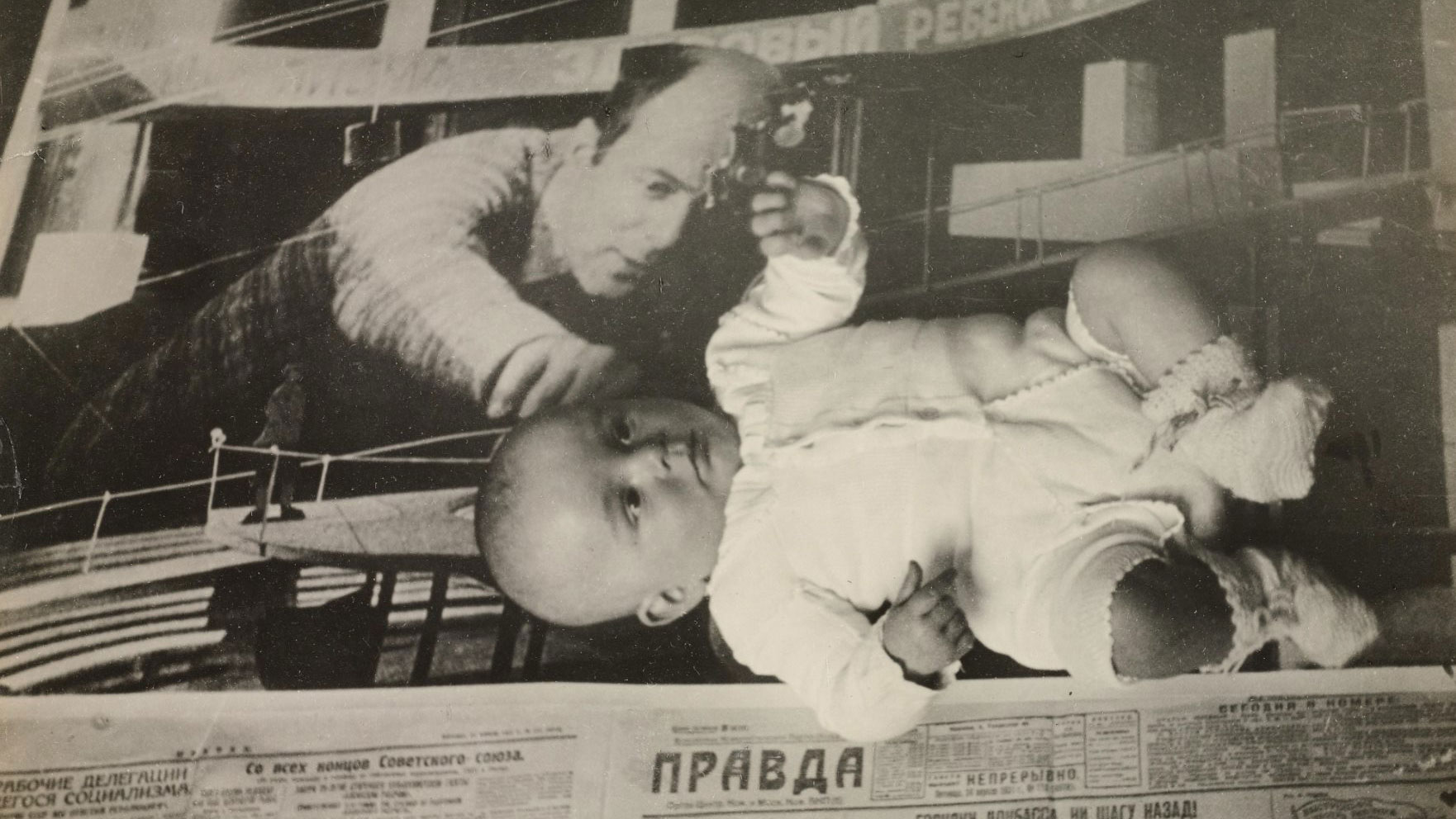 A vintage black and white Russian ad in a newspaper of a balding man reaching across a mechanical item. A baby wearing a sweater and booties is laying on top of the paper.  