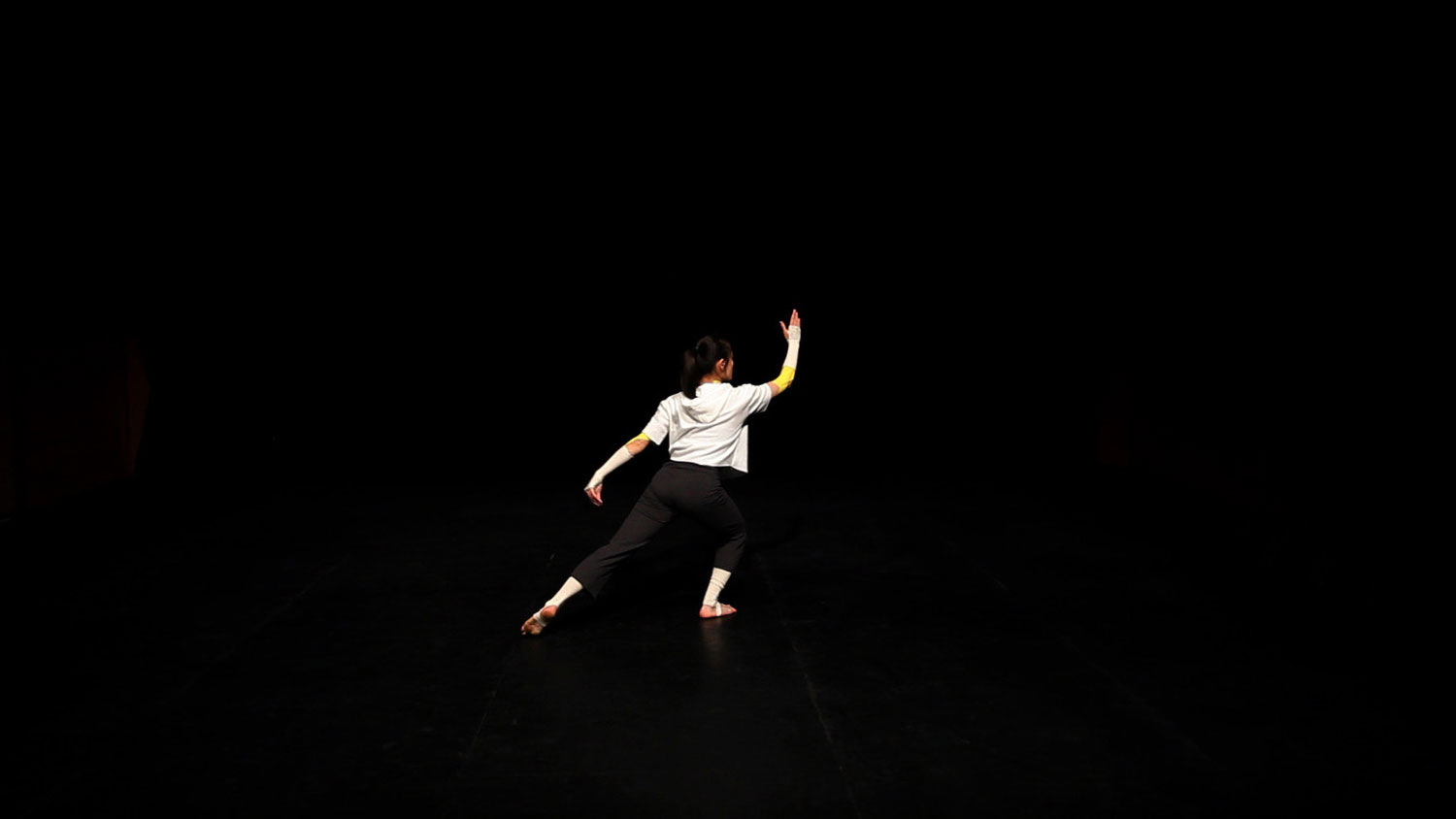 A woman wearing a white top and black bottoms on a black stage facing forward with her arm raised in motion. 