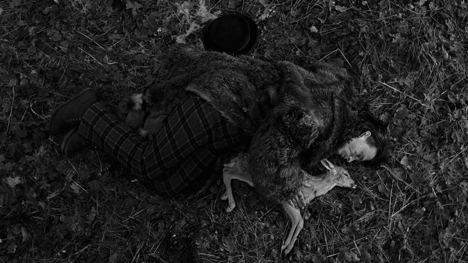 A man wearing a fur coat and tartan pants laying on the ground next to a deceased fawn in a cuddling position. 