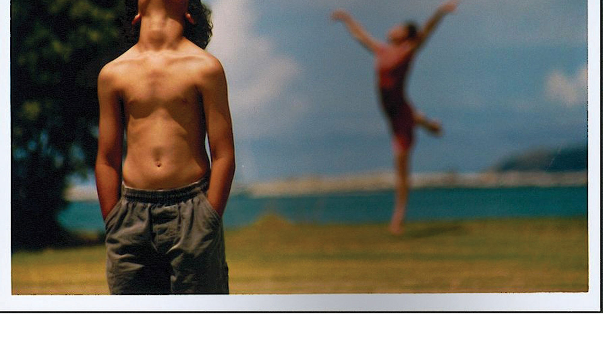 A young boy standing on a beach with tanned skin wearing a gray bathing suit looking up with his head cut out of the image. Behind him another person can be seen leaping. 