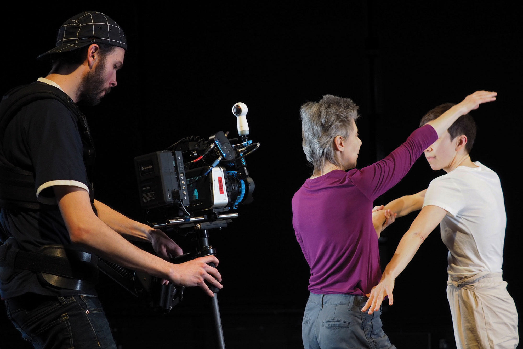 Two female dancers with short hair, one wearing purple the other wearing white and a steadicam operator shooting in a black box studio. 