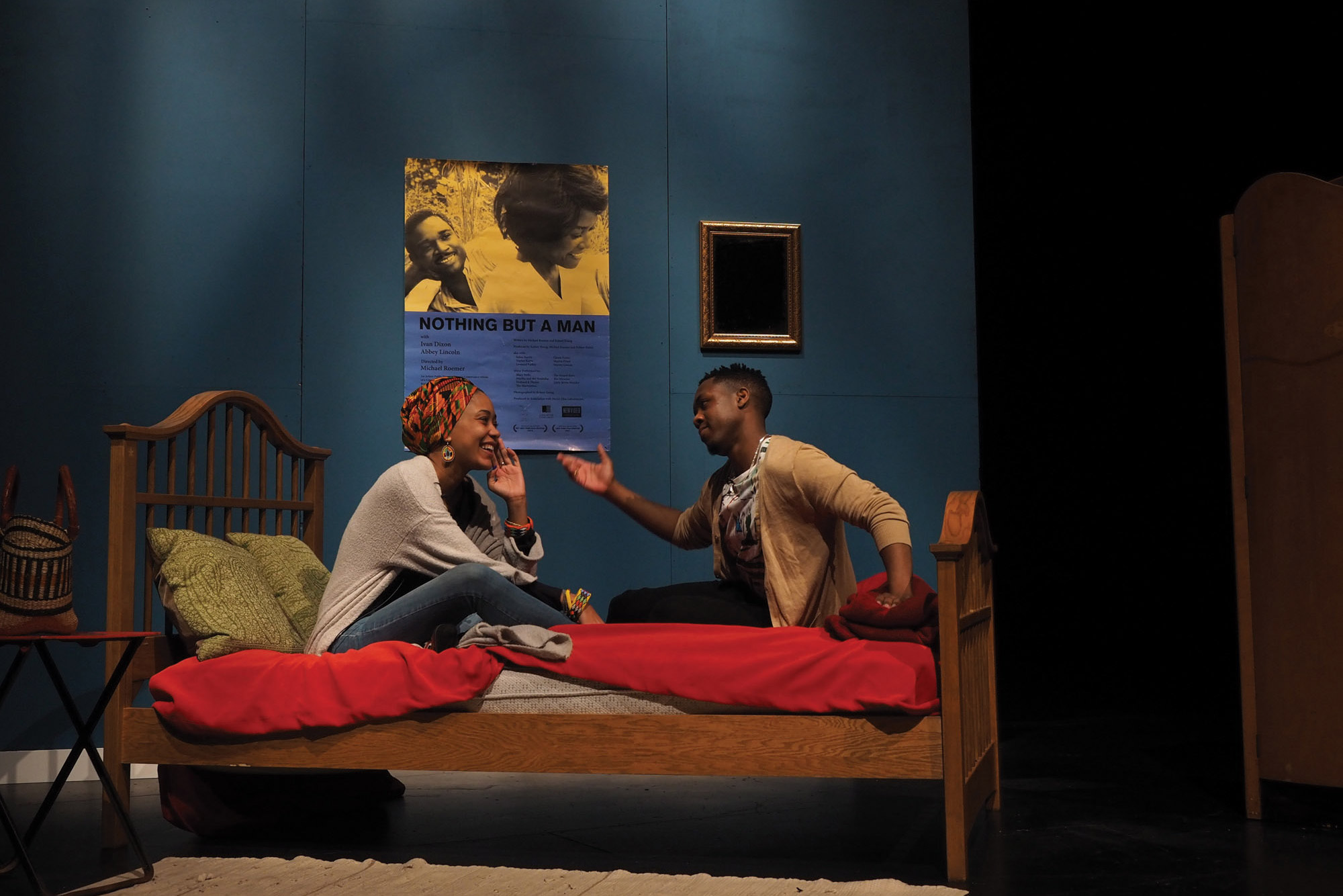 A Black woman and a Black man in a blue room sitting on a bed with red sheets in animated joyous conversation. 