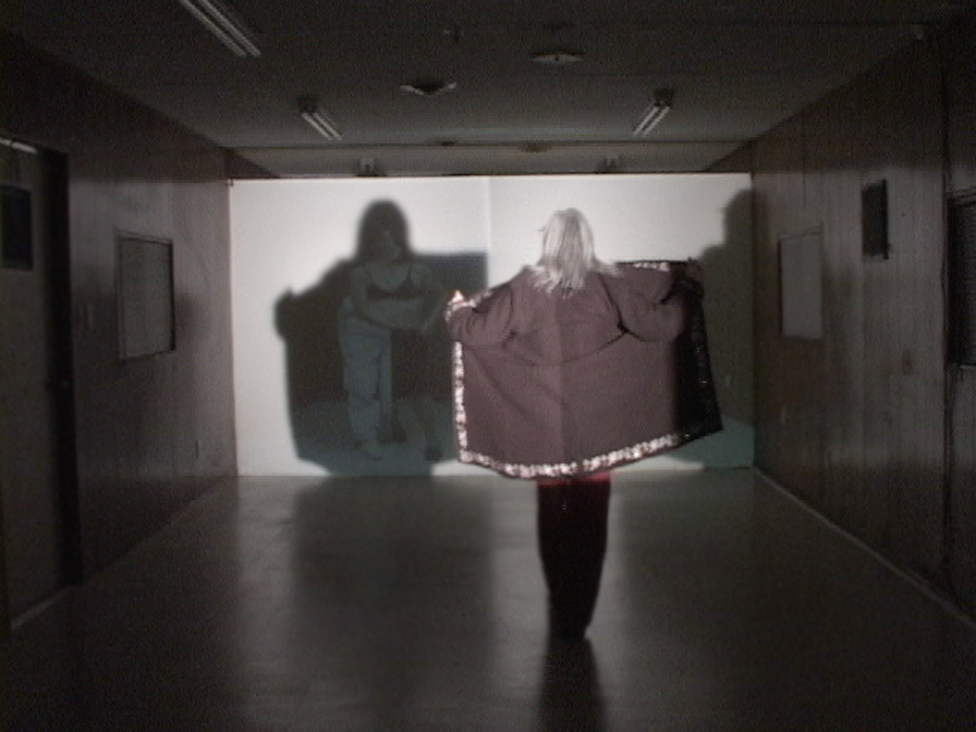 a woman in a gray room creates a shadow by holding open her jacket against a screen. 