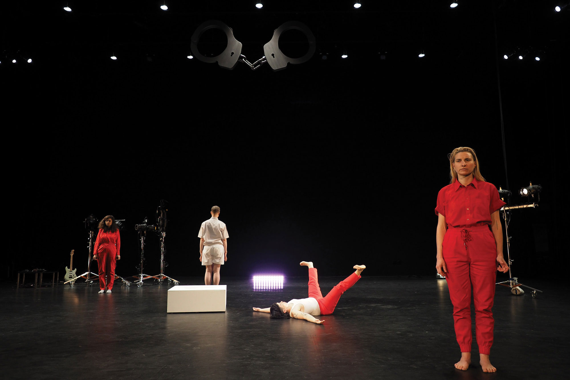 A woman wearing a red jumpsuit standing in front of three dancers wearing red and white in various poses in a black box studio. 