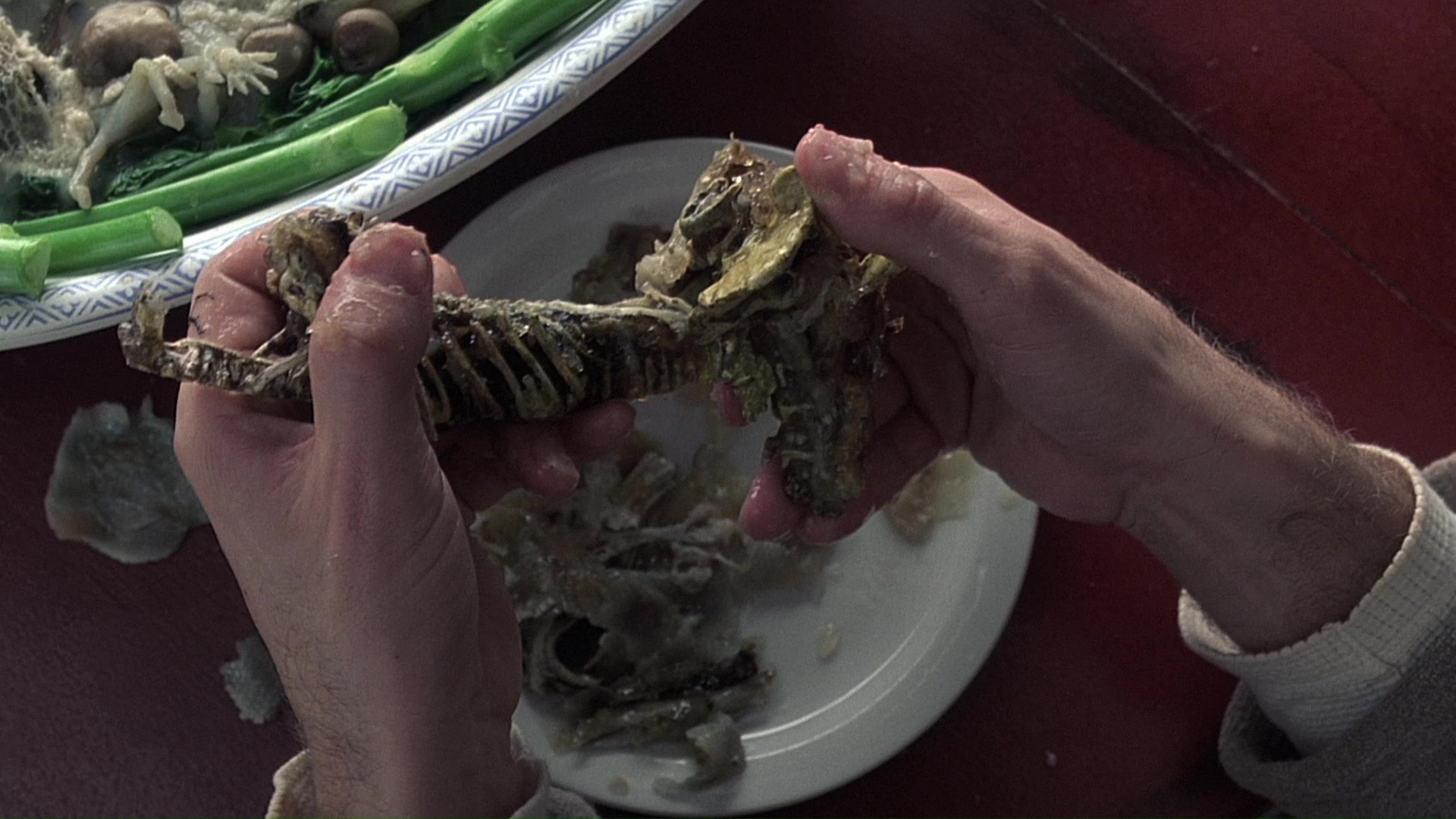 Two white hands holding a small carcass over a dinner plate as pieces of meat fall to plate below. A serving platted of other meats on a bed of greens is in the top corner. 