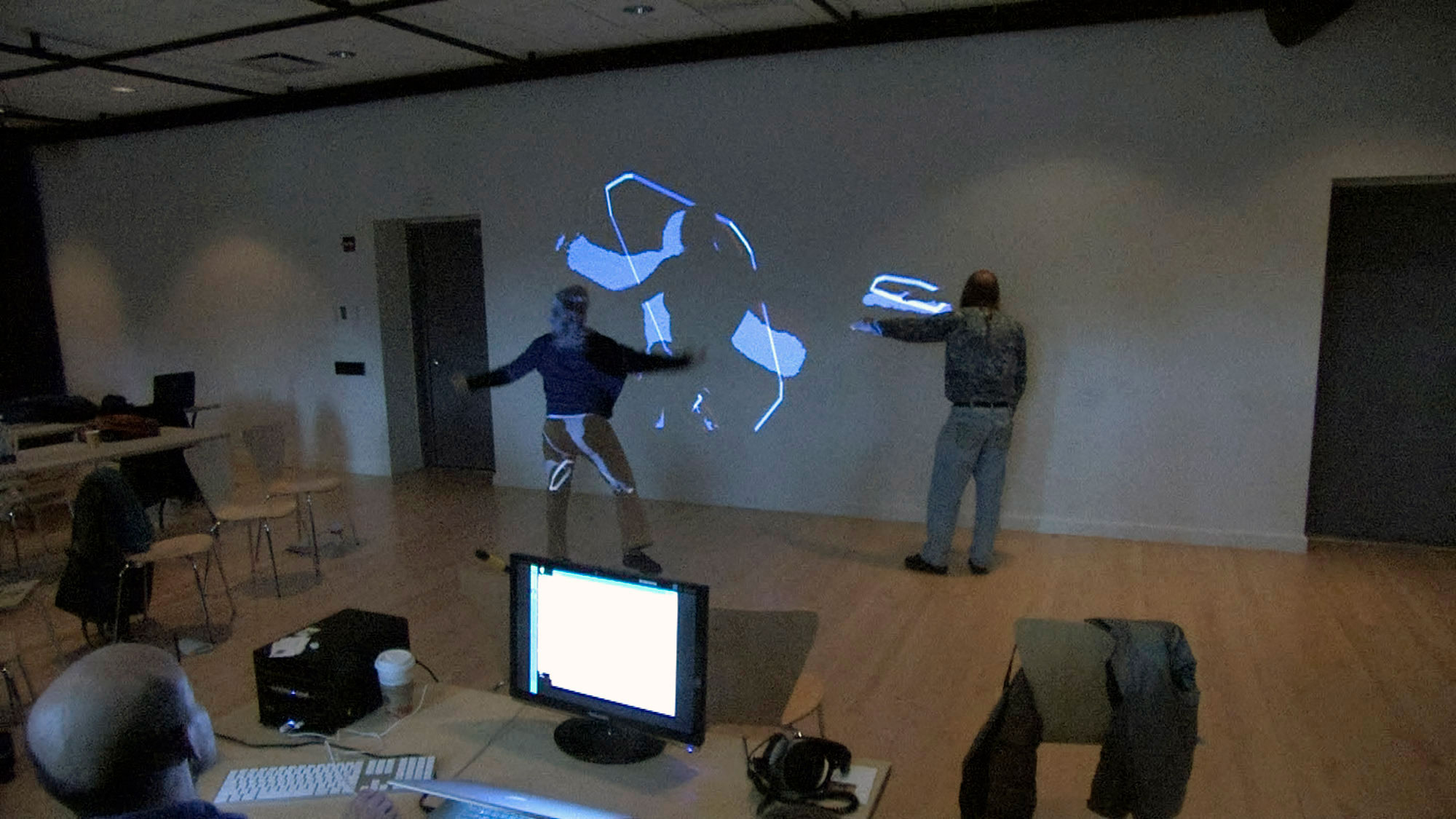 Two people with backs to the viewer interacting with a wall projection controlled by a bald man sitting at a computer in the foreground. 