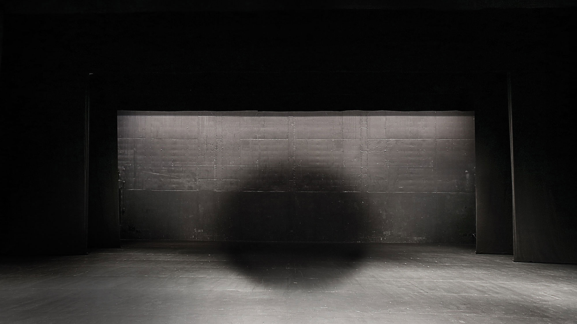 A black circle of filtered light projected between the wall and floor of a black box studio. 