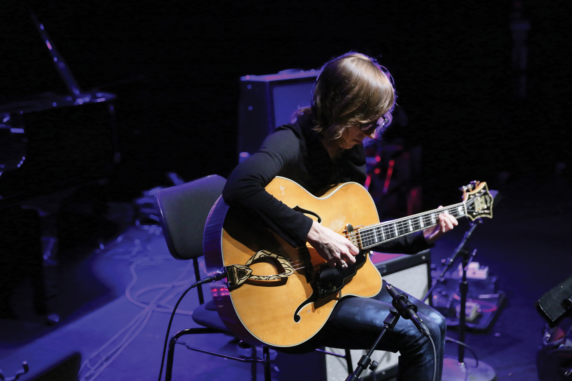 A woman playing an amplified acoustic guitar washed in purple light. 