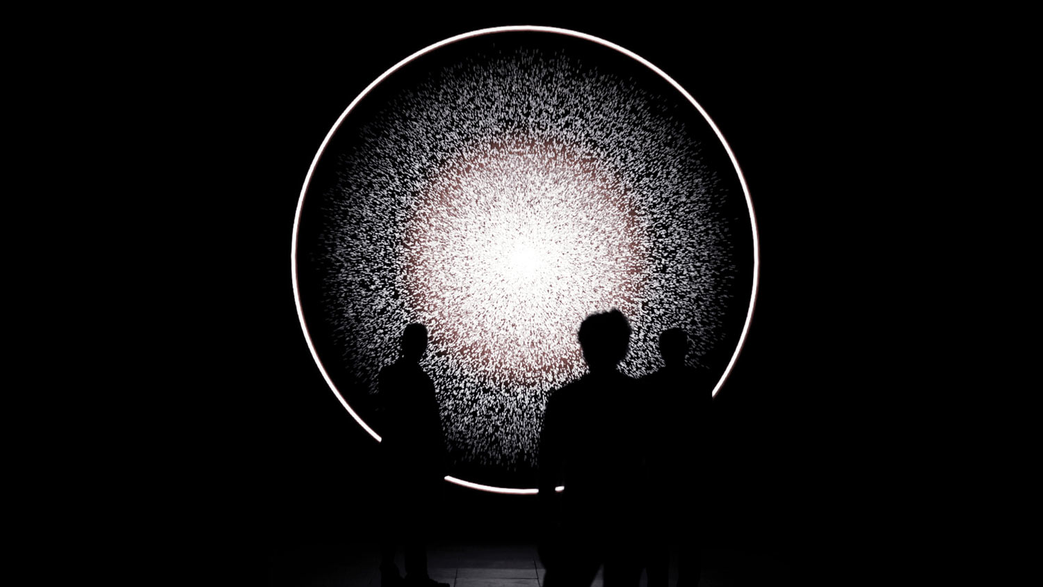 A ring of light surrounding spore like dots of light in a dark room. A small group of three people silhouetted looks on 