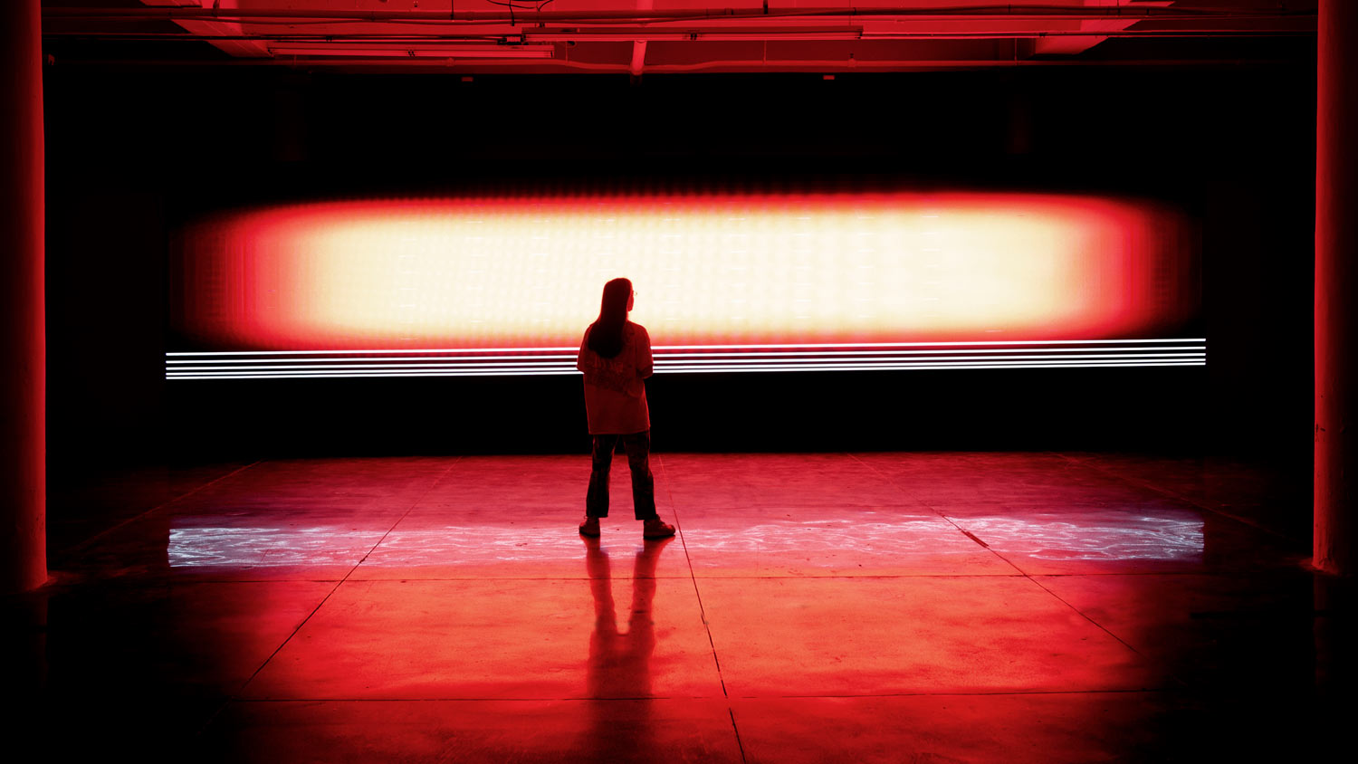 A woman stand with back to the viewer in front of a strip of red light in a dark theater.  