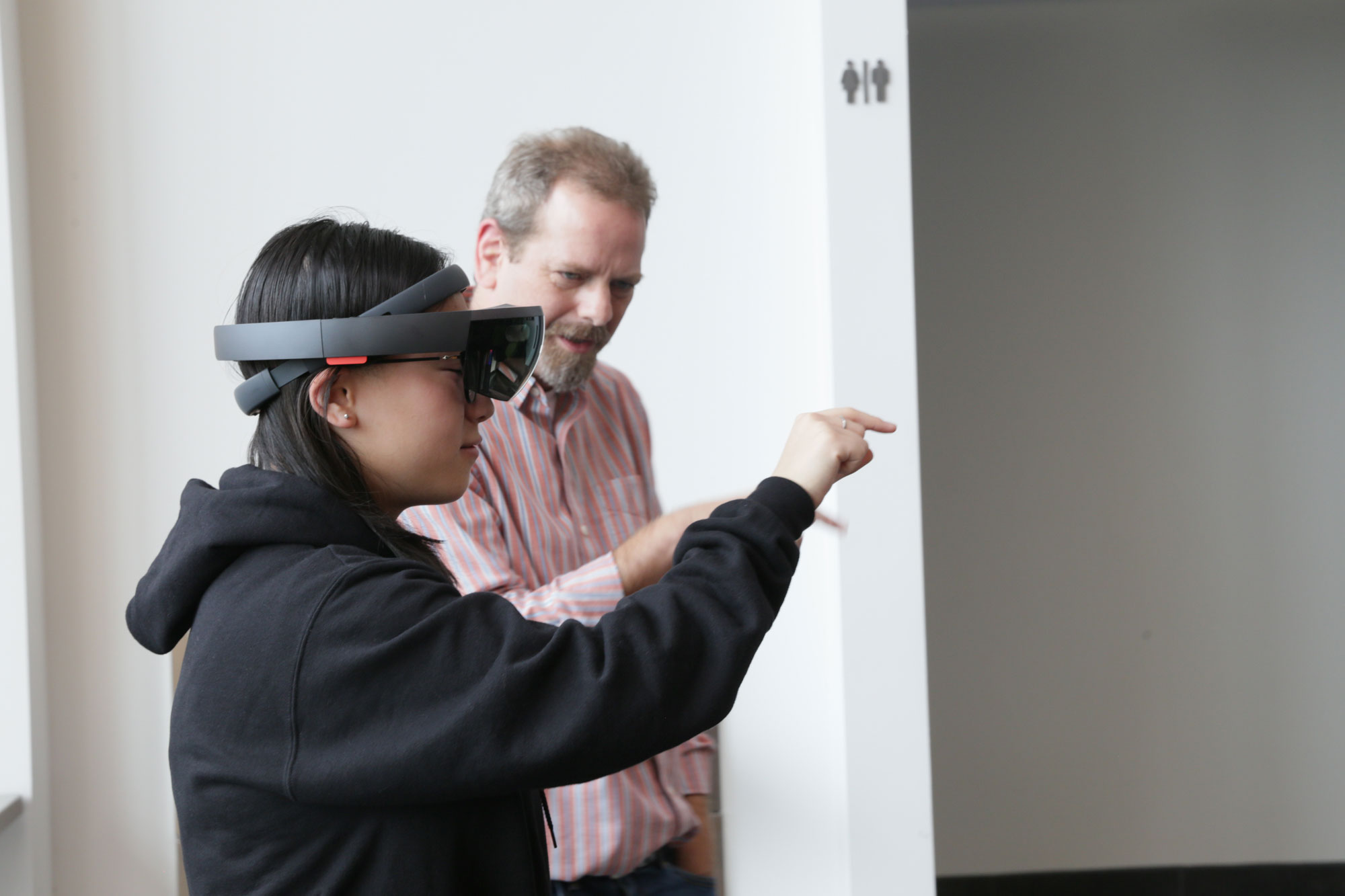 A woman wearing VR goggles pointing while being facilitated by Dave Bebb. 