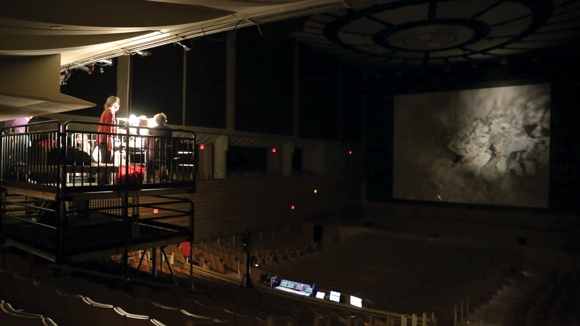 Two people on scaffolding projecting an image of a gray ink blob onto a large screen on the concert hall stage.  