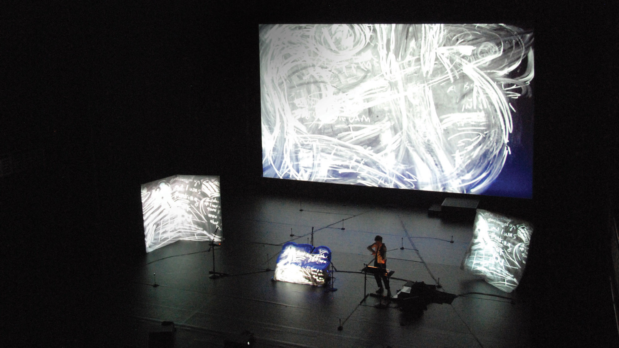 Laurie Anderson in lecture surrounded by screens of various sizes projecting images of chalk swirls. 