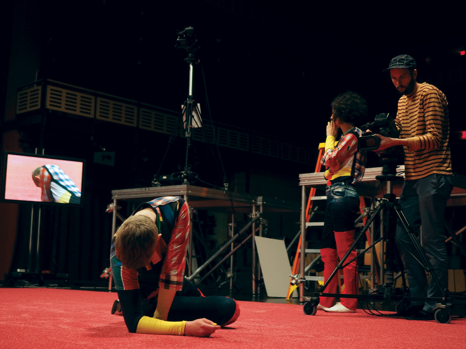 A dancer wearing a colorful costume laying on a red carpet while being filmed as Maria Hassabi looks at the video feed on a screen, contemplating. 