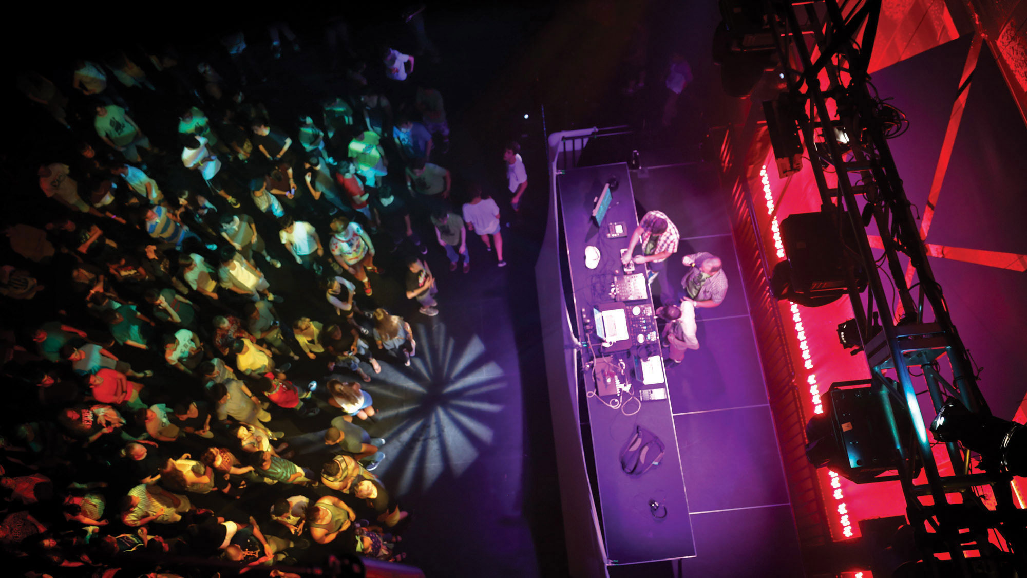 An aerial shot of three men DJing on stage performing for a crowd in a wash of rainbow party lights. 