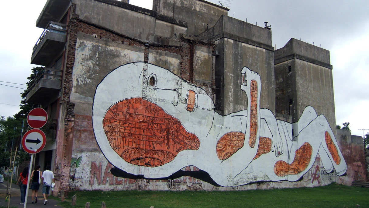 Graffiti of a human laying on their back holding a pill up to take it on a decrepit industrial building.