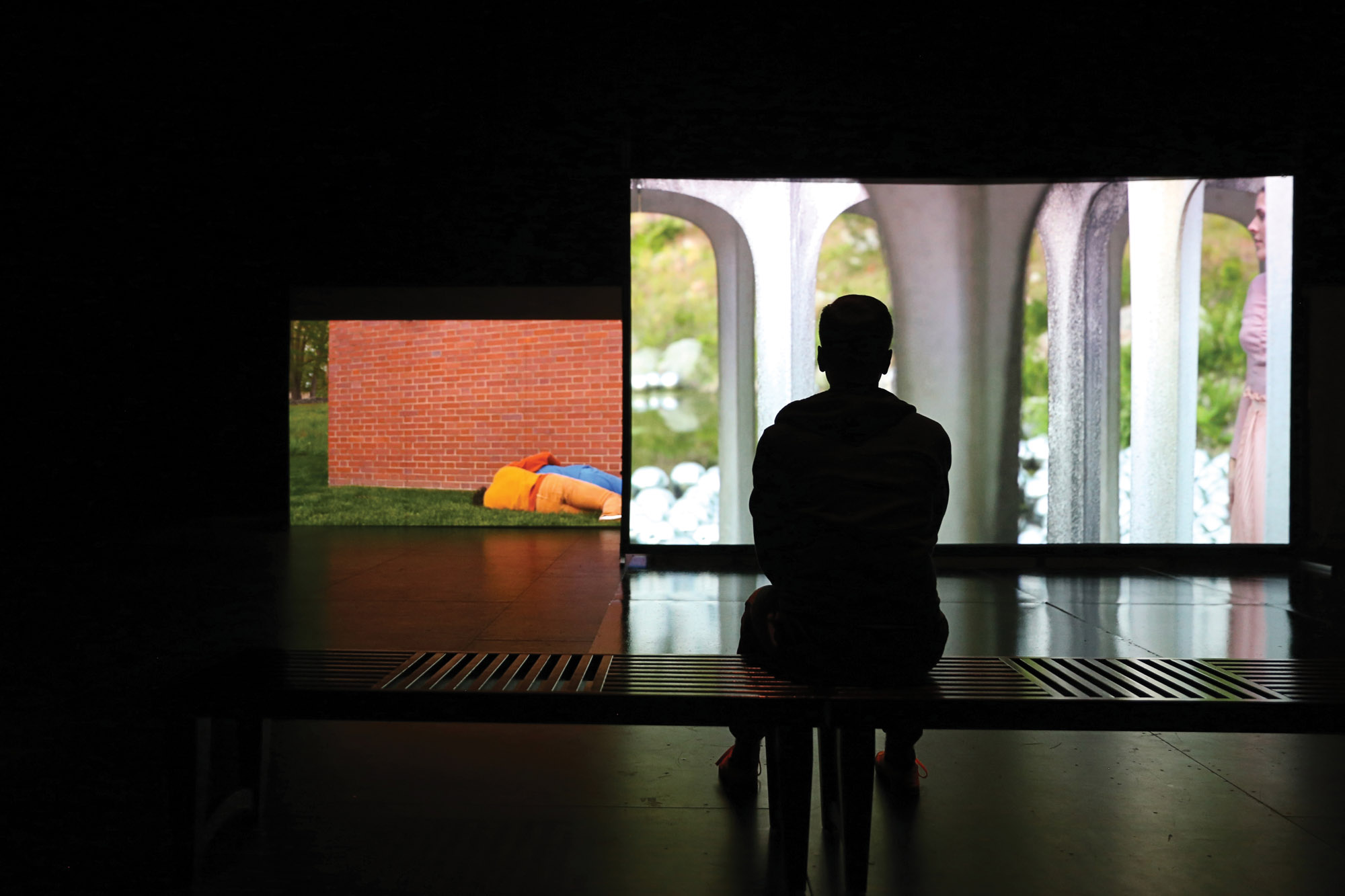 a silhouetted man sitting on a bench viewing two large video screens with projections of a brick wall concrete arches. 