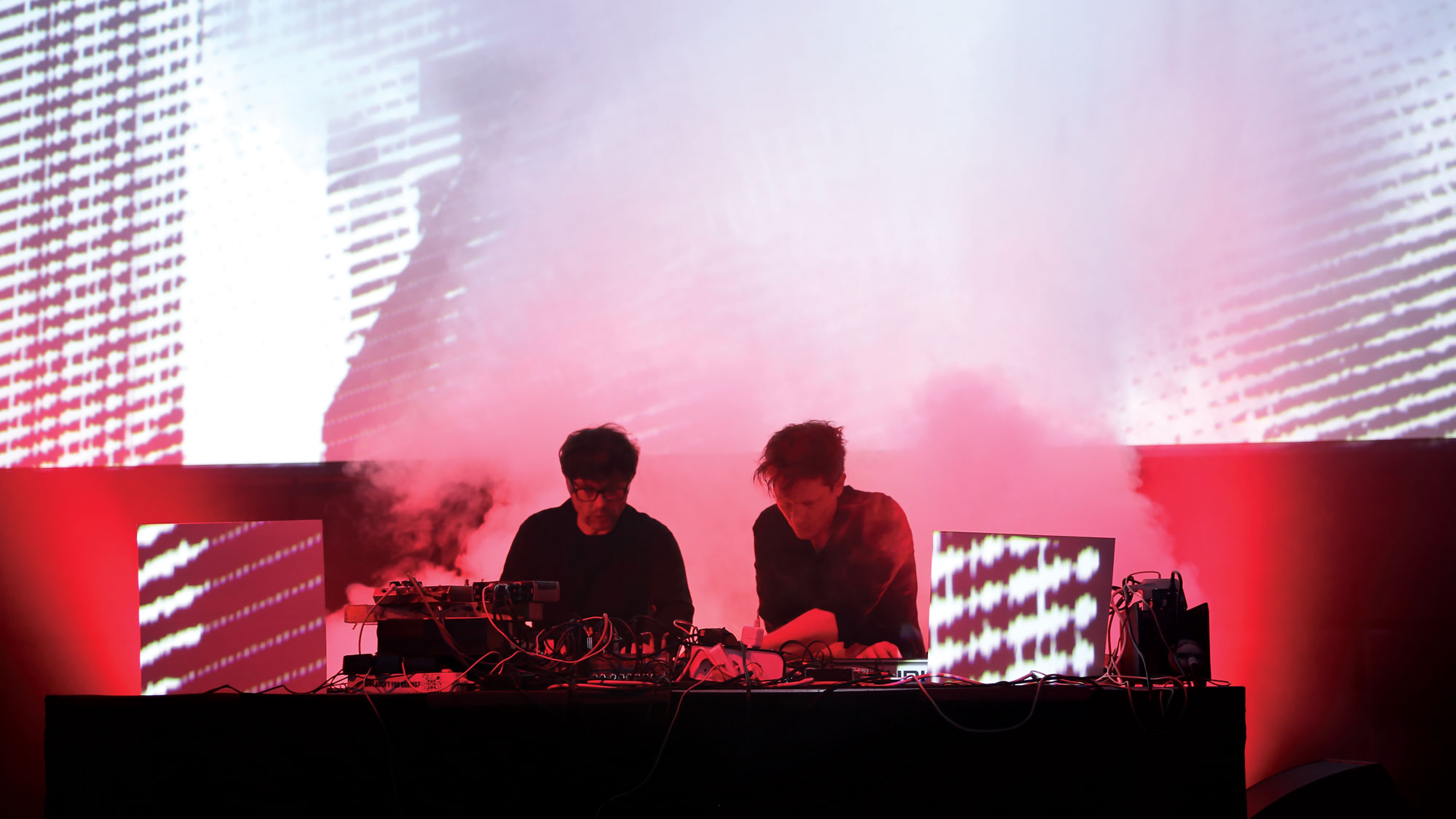 Two men DJing on a stage in front of was of red light and theatrical smoke. 
