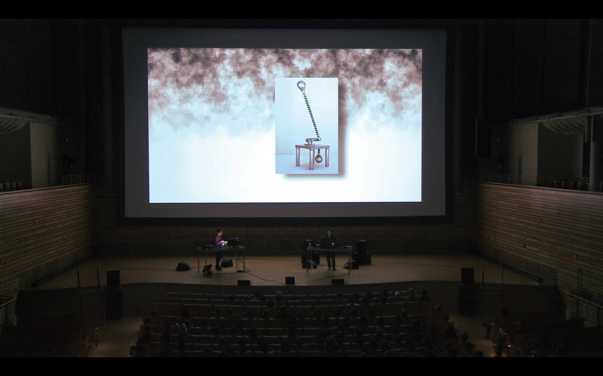 Two people behind desks and in discussion on stage infant of a large screen projecting the image of an abstract sculpture, resembling a low table with a long handle. 