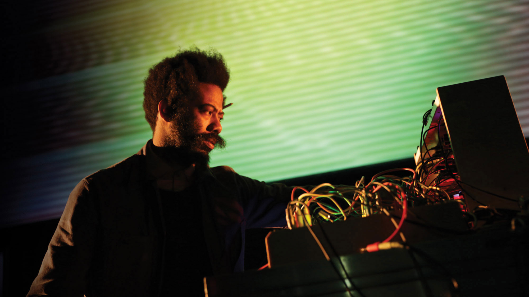 A Black man with a prominent mustache in front of a green and yellow projection examining various colored wires. 