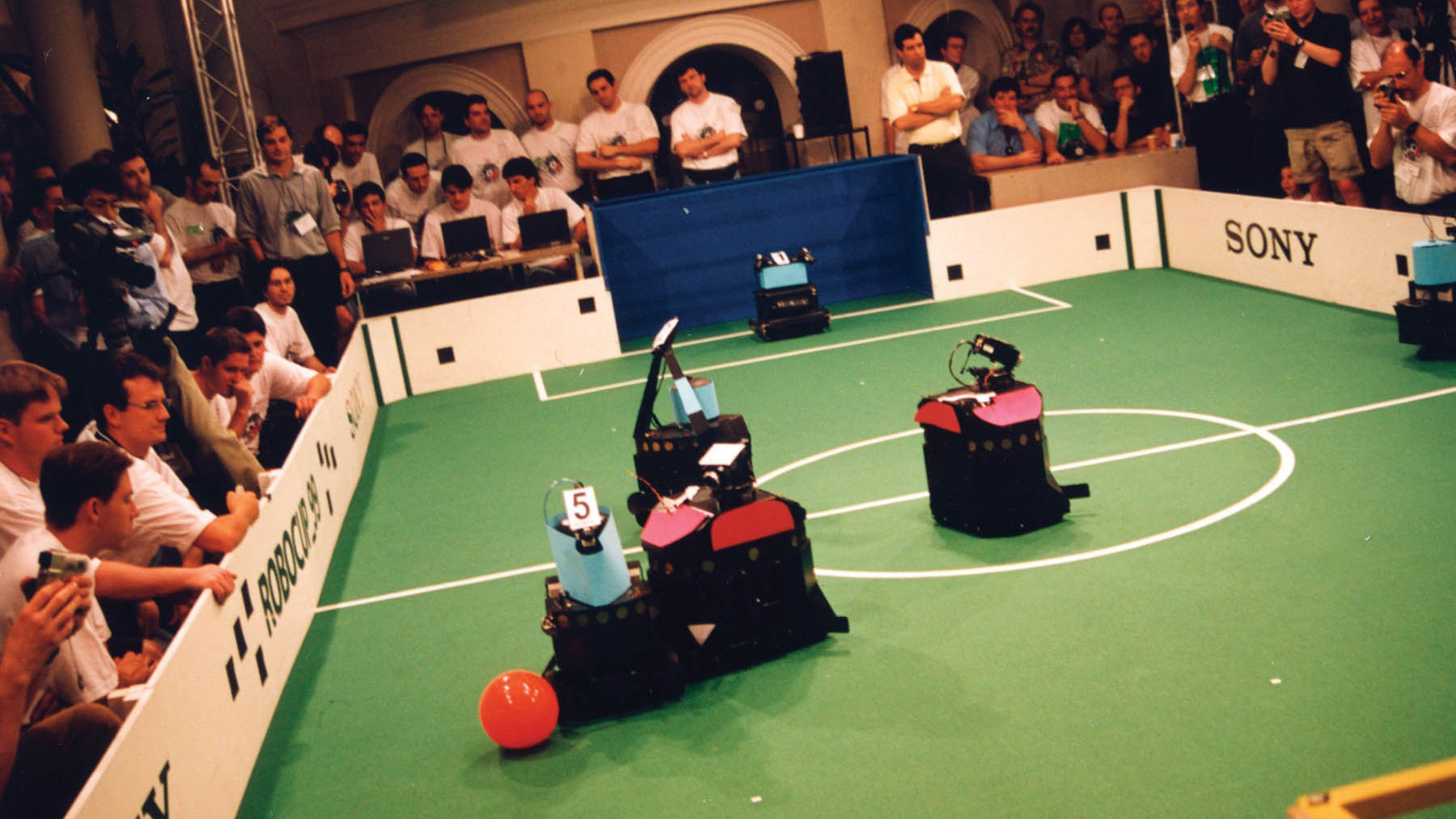 Six robots wearing blue or red on a pitch playing a form of soccer with a red ball surrounded by men controlling them. 