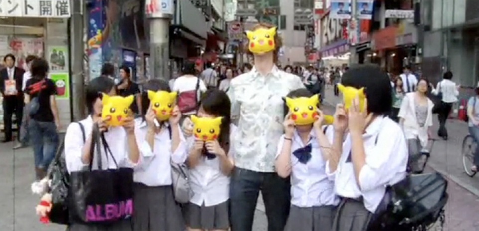 A tall white man with a group of five asian woman posing with Pikachu masks on a bustling city street. 