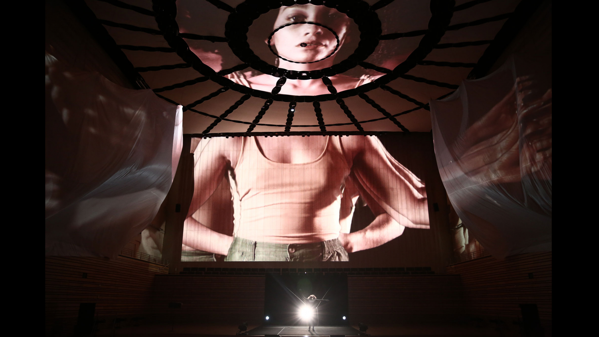 Projections of a female on the concert hall stage. Her head on the ceiling, torso on the back wall and arms outstretched on flowing fabric hung from the sides of the stage. 