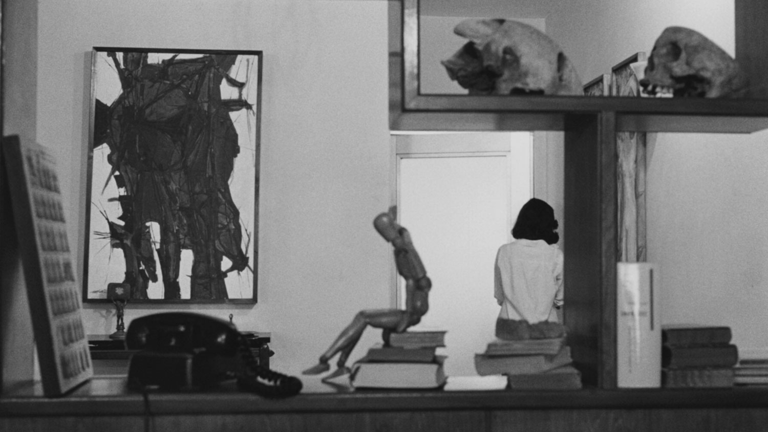 A table parallel to the view of the camera with books, a wooden artists dummy, and a rotary phone. A neatly decorated apartment with an AbEx painting can be seen in the background. A woman stand with back to the viewer leaning against the wall. 