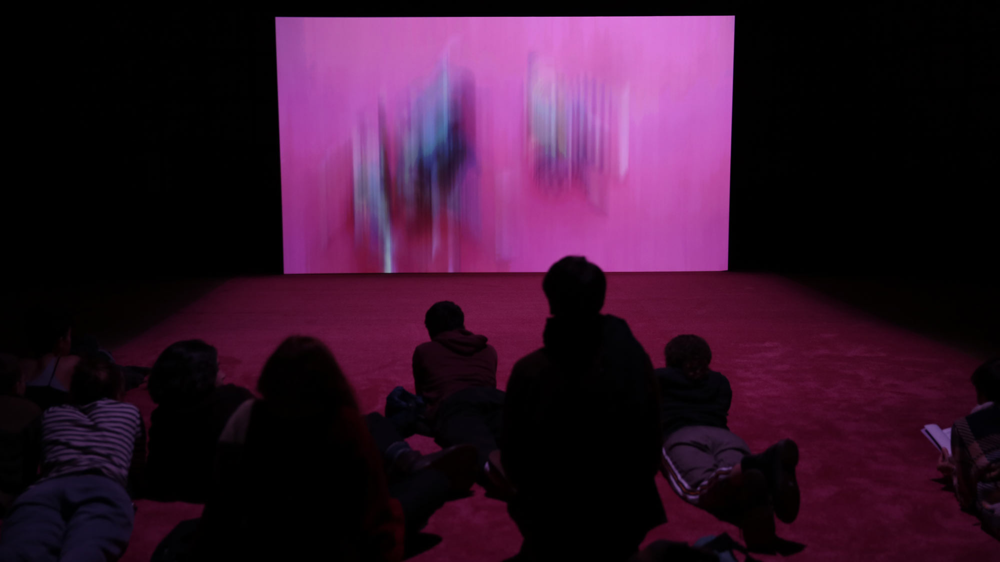 A small silhouetted audience laying on a pink rug watching a projection of pink brush strokes in a dark room. 