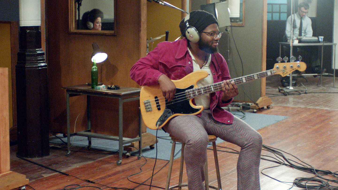 A Black man wearing a beanie and dressed in 1970's fashion plays the bass in the middle of a 70's era recoding studio. 